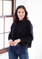 A woman with dark hair is wearing a black long-sleeve knit top and blue jeans, standing in a brightly lit room with a window in the background.