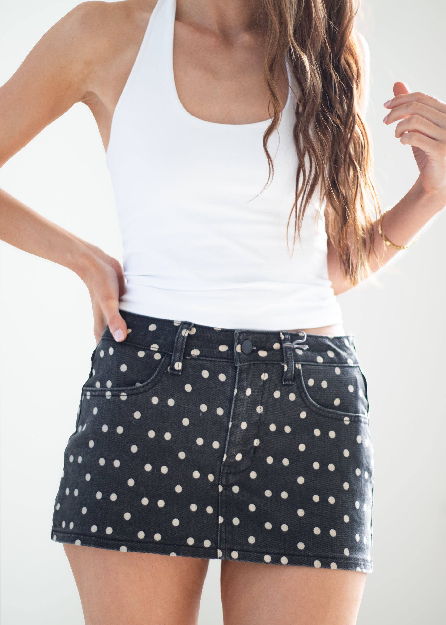 A woman wearing a white ribbed tank top and a black denim mini skirt with a white polka dot pattern, photographed against a clean white background.