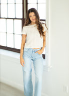 Woman wearing a white top and light blue jeans standing in a bright room with large windows.