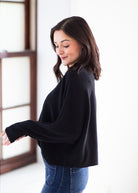 A woman wearing a black textured knit long-sleeve sweater and blue jeans, standing in a bright indoor setting.