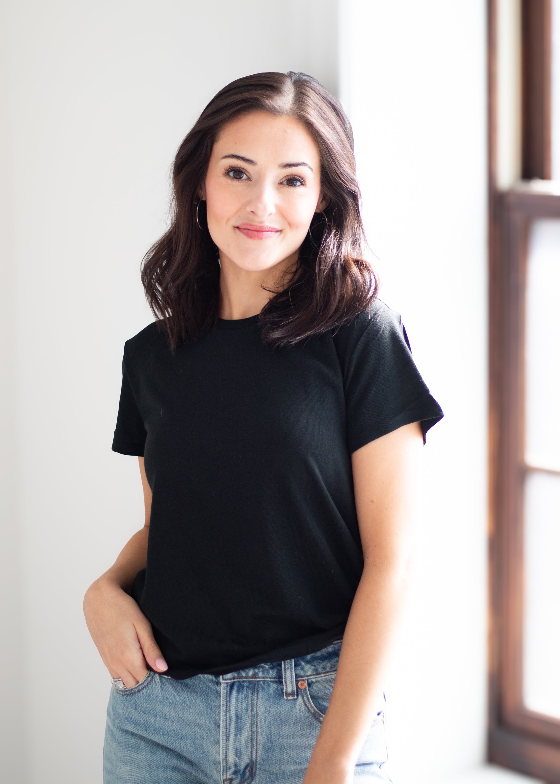 A woman with shoulder-length brown hair wearing a plain black short-sleeve crewneck t-shirt and light-wash blue jeans, standing in a brightly lit room next to a window.