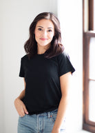 A woman with shoulder-length brown hair wearing a plain black short-sleeve crewneck t-shirt and light-wash blue jeans, standing in a brightly lit room next to a window.