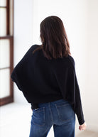 A woman viewed from behind wearing a black knit sweater and blue jeans in a bright indoor setting.