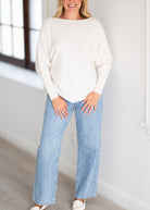 A woman wearing a cream-colored boatneck knit sweater and light-wash wide-leg jeans, standing in a bright room with a window.