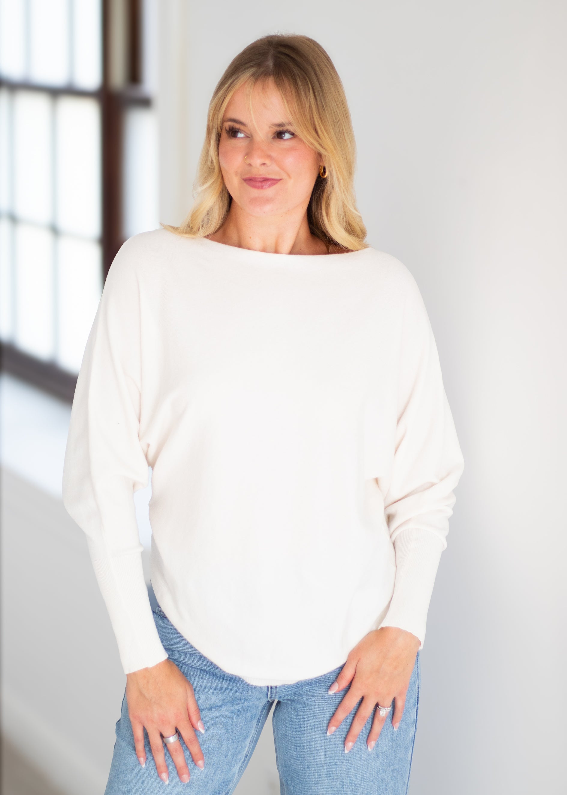 A woman with blonde hair wearing a cream-colored, long-sleeve boat-neck sweater and light blue jeans, standing in a brightly lit room.