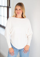 A woman with blonde hair wearing a cream-colored, long-sleeve boat-neck sweater and light blue jeans, standing in a brightly lit room.