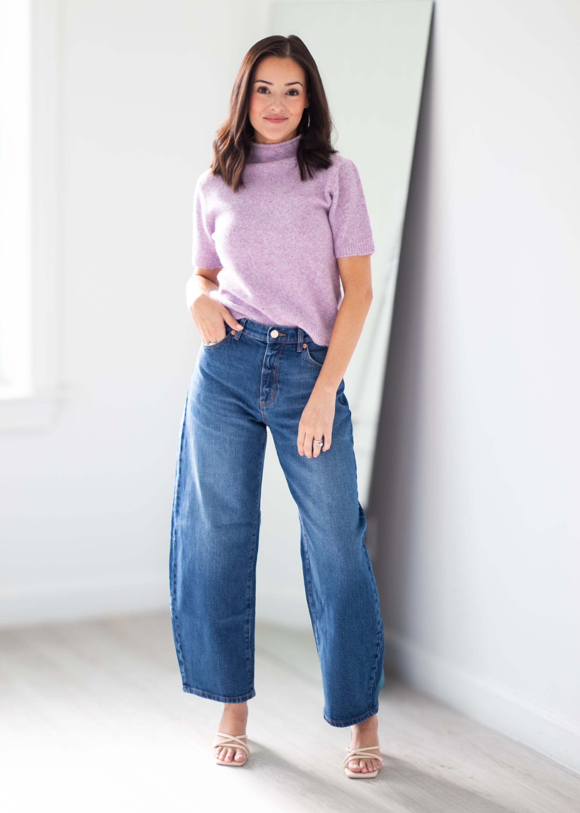 A woman modeling a lavender short-sleeve knit mock neck top and high-waisted, wide-leg blue jeans, standing in a bright, minimalist room with white walls and light wood flooring.