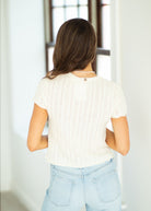Woman wearing a white knit top and light blue jeans in a bright room.