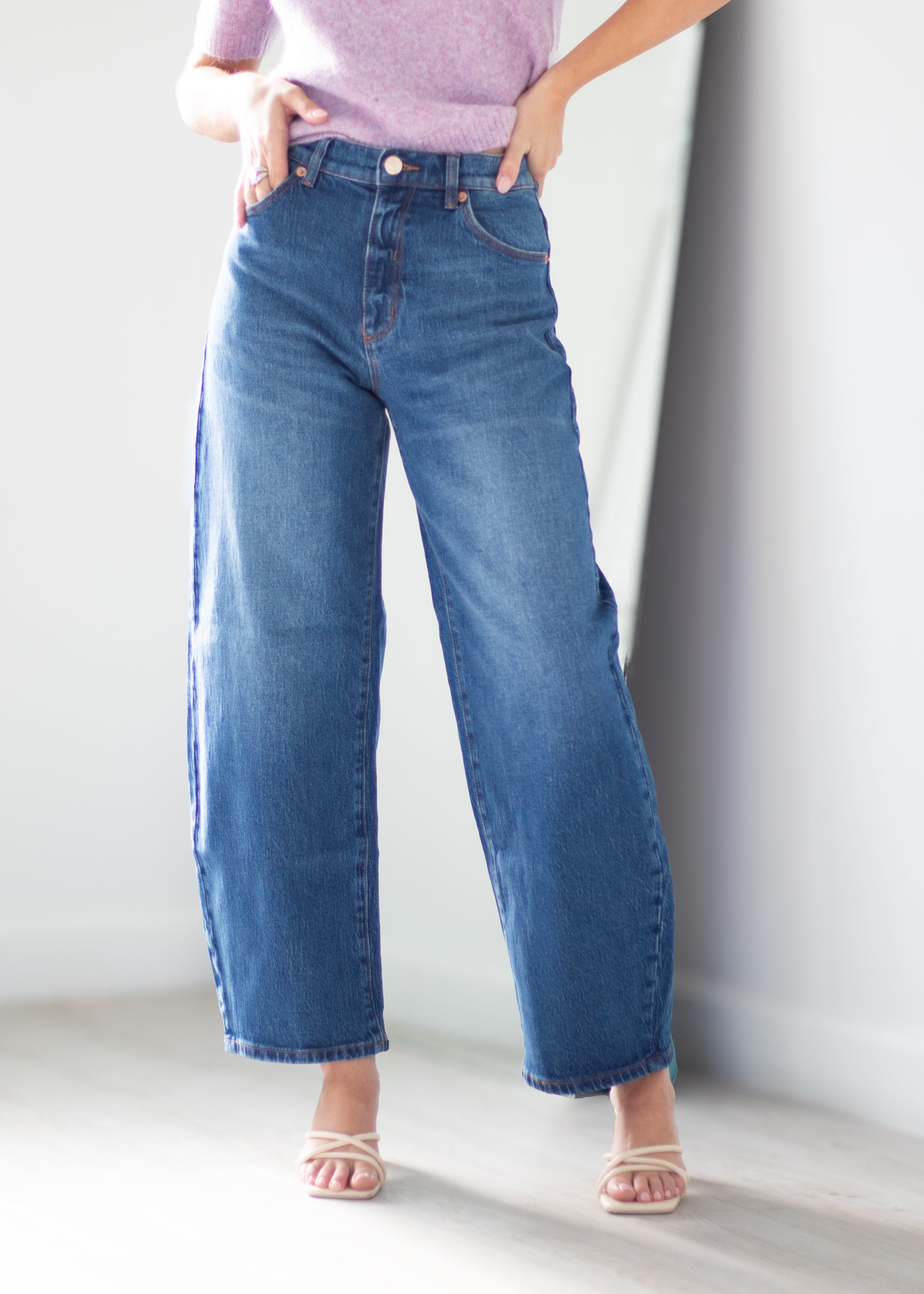A person wearing medium-wash blue wide-leg cropped jeans with side pockets, paired with a light purple top and beige strappy heels, standing against a white wall.