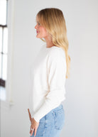 A woman with blonde hair is shown in profile wearing a cream-colored long-sleeve knit sweater and light blue jeans against a bright white background.
