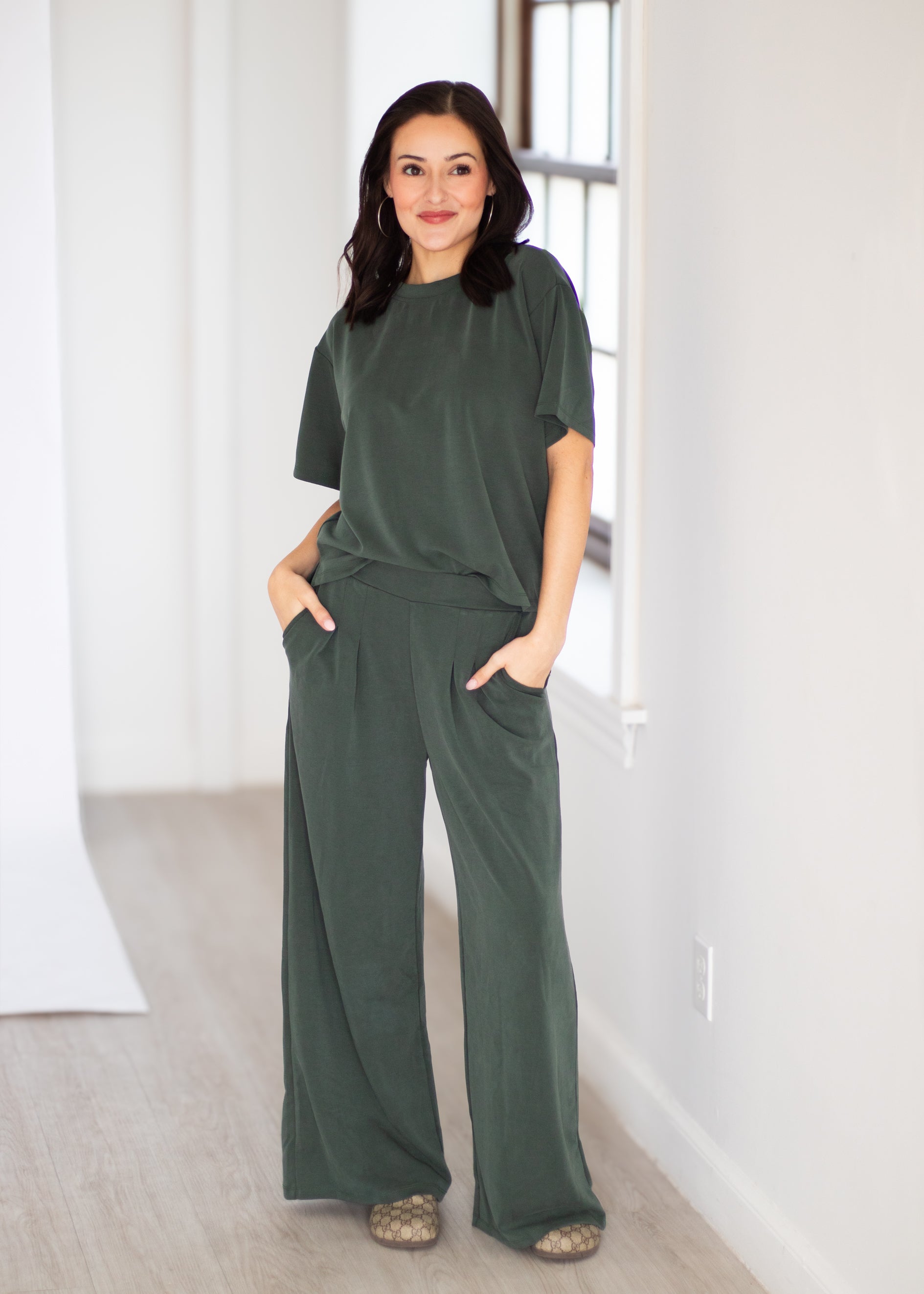 A woman modeling a dark olive green two-piece lounge set consisting of a short-sleeve crewneck top and matching wide-leg pants, standing in a bright, minimalist room with white walls and light wood flooring.