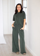 A woman modeling a dark olive green two-piece lounge set consisting of a short-sleeve crewneck top and matching wide-leg pants, standing in a bright, minimalist room with white walls and light wood flooring.
