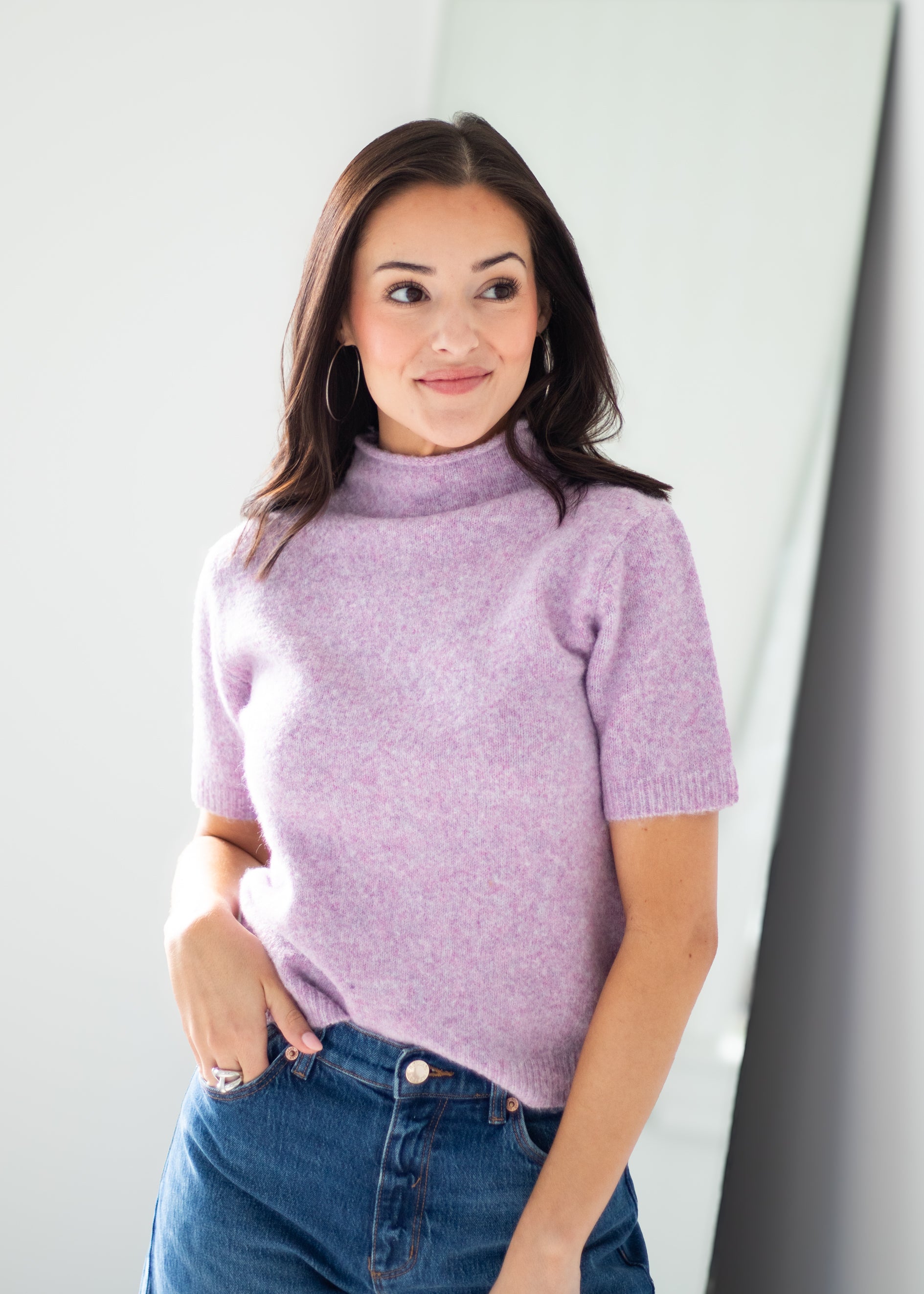 A woman wearing a short-sleeve knit mock neck sweater in a light lavender purple, paired with blue jeans, standing against a minimalist light gray background.
