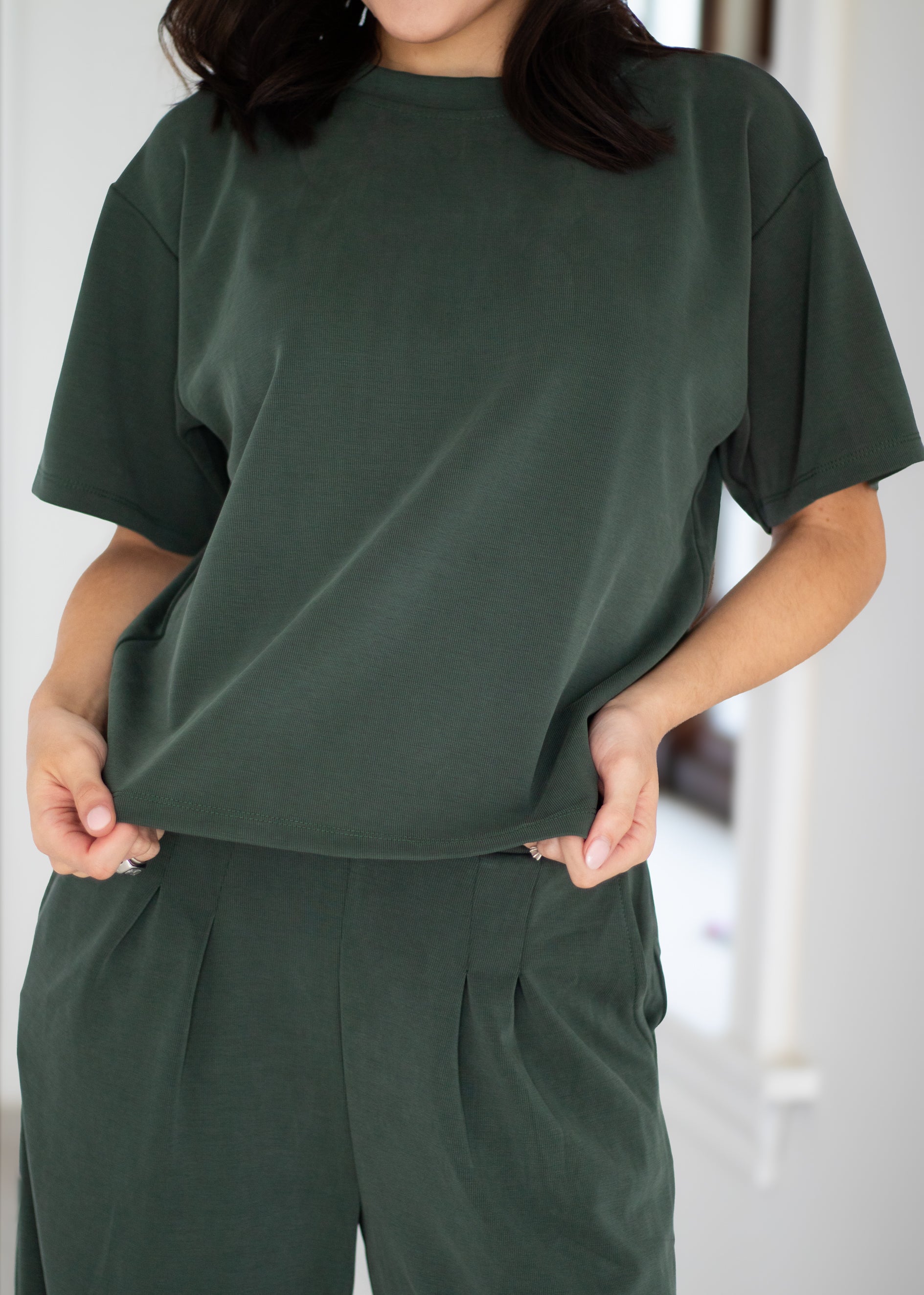 A person wearing an oversized dark green short-sleeve top and matching dark green pleated pants, shown from the neck down in a bright indoor setting.