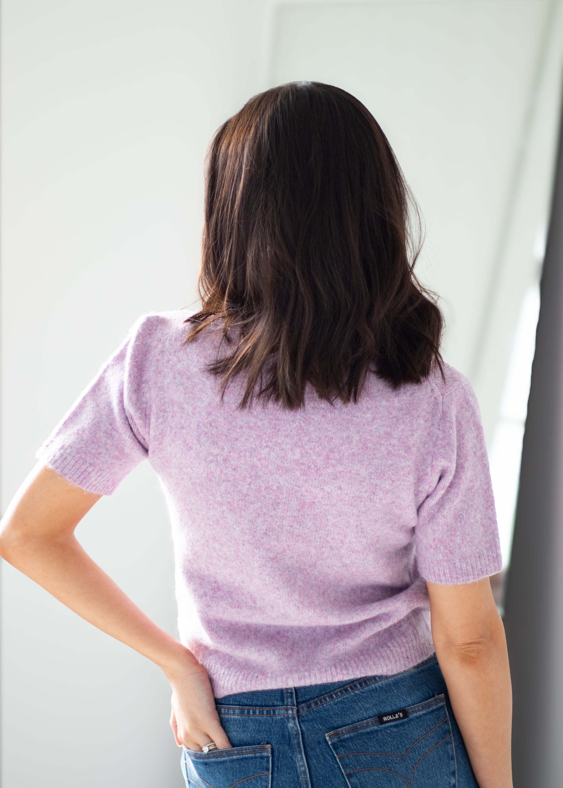 A woman viewed from behind wearing a short-sleeve lavender knit sweater and blue jeans, standing against a bright white wall.