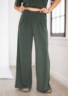 A person wearing dark olive green wide-leg pleated pants, paired with a matching dark green cropped top and patterned flat shoes, standing in a bright indoor room.