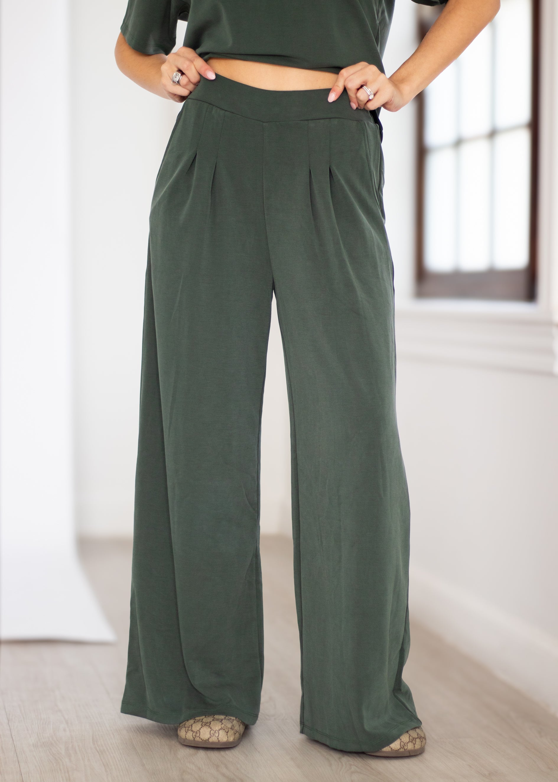 A person wearing dark olive green wide-leg pleated pants, paired with a matching dark green cropped top and patterned flat shoes, standing in a bright indoor room.