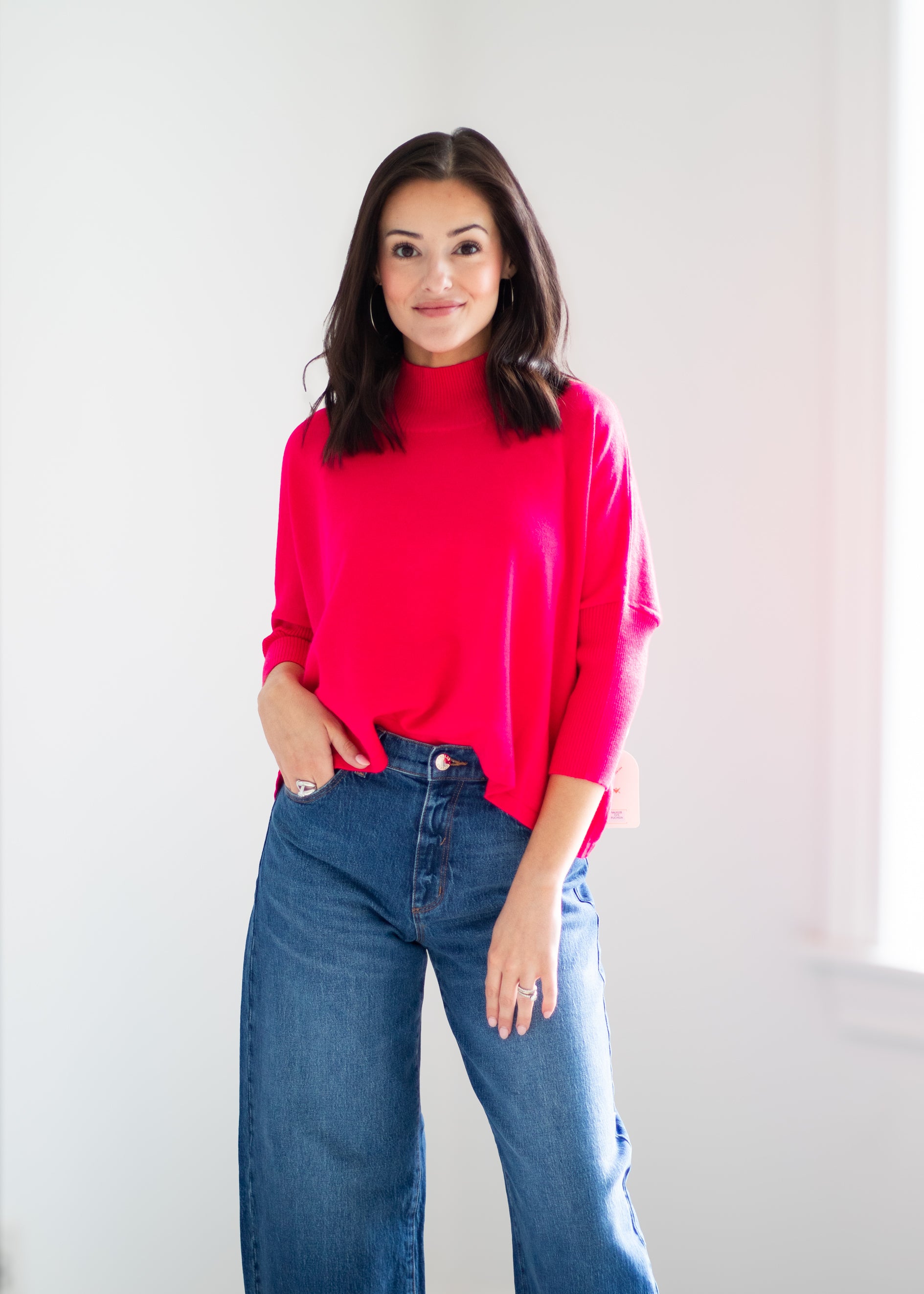 A woman wearing a bright pink mock-neck long-sleeve top and blue jeans, standing against a plain white wall.