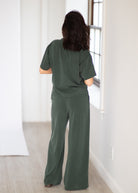 A woman viewed from behind wearing a matching dark green short-sleeve top and wide-leg pants set in a bright, minimalist room with light wood floors and a window.