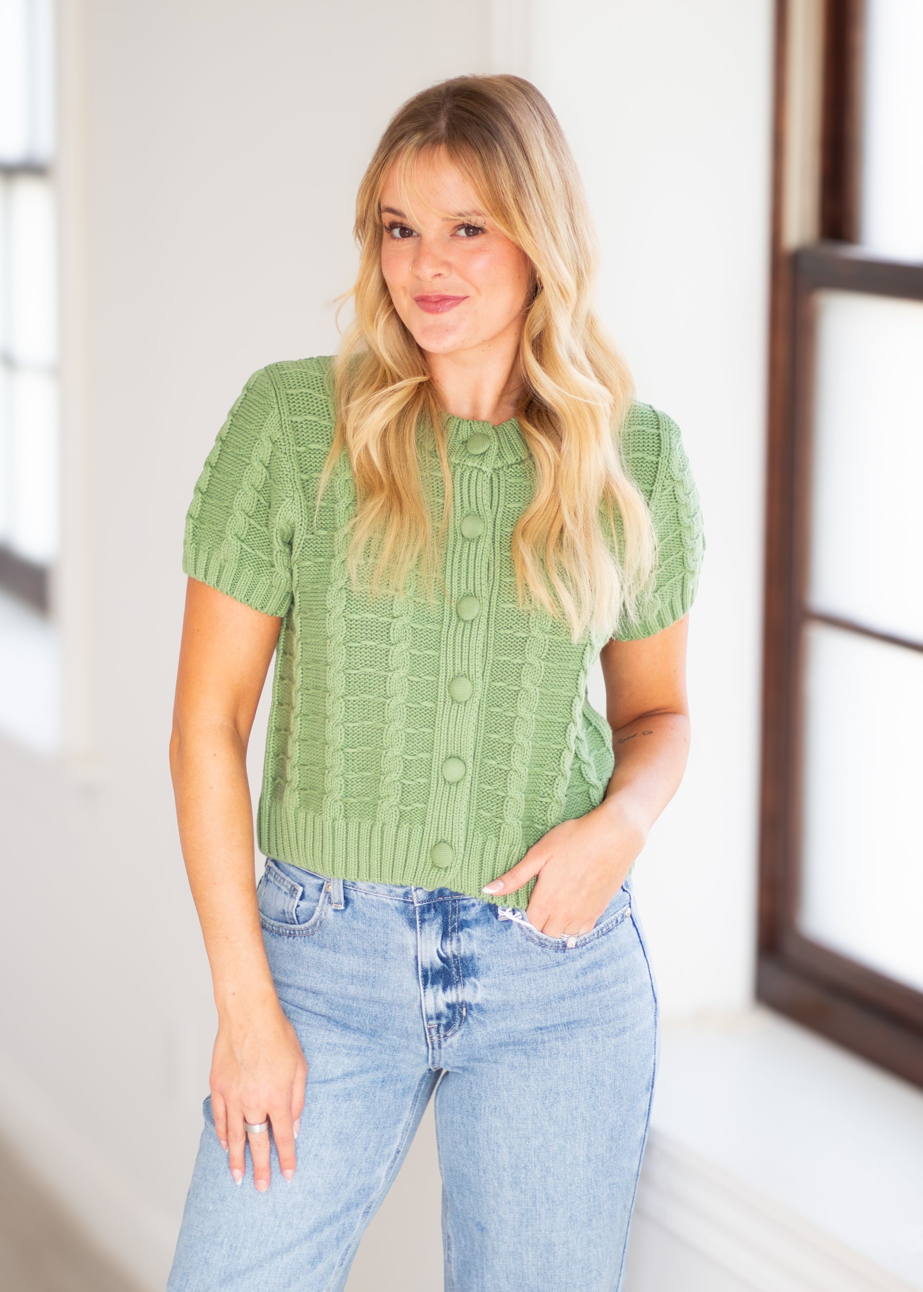 A woman with blonde hair wearing a sage green short-sleeve button-up knit cardigan and light wash blue jeans, standing in a brightly lit room with white walls and window frames.