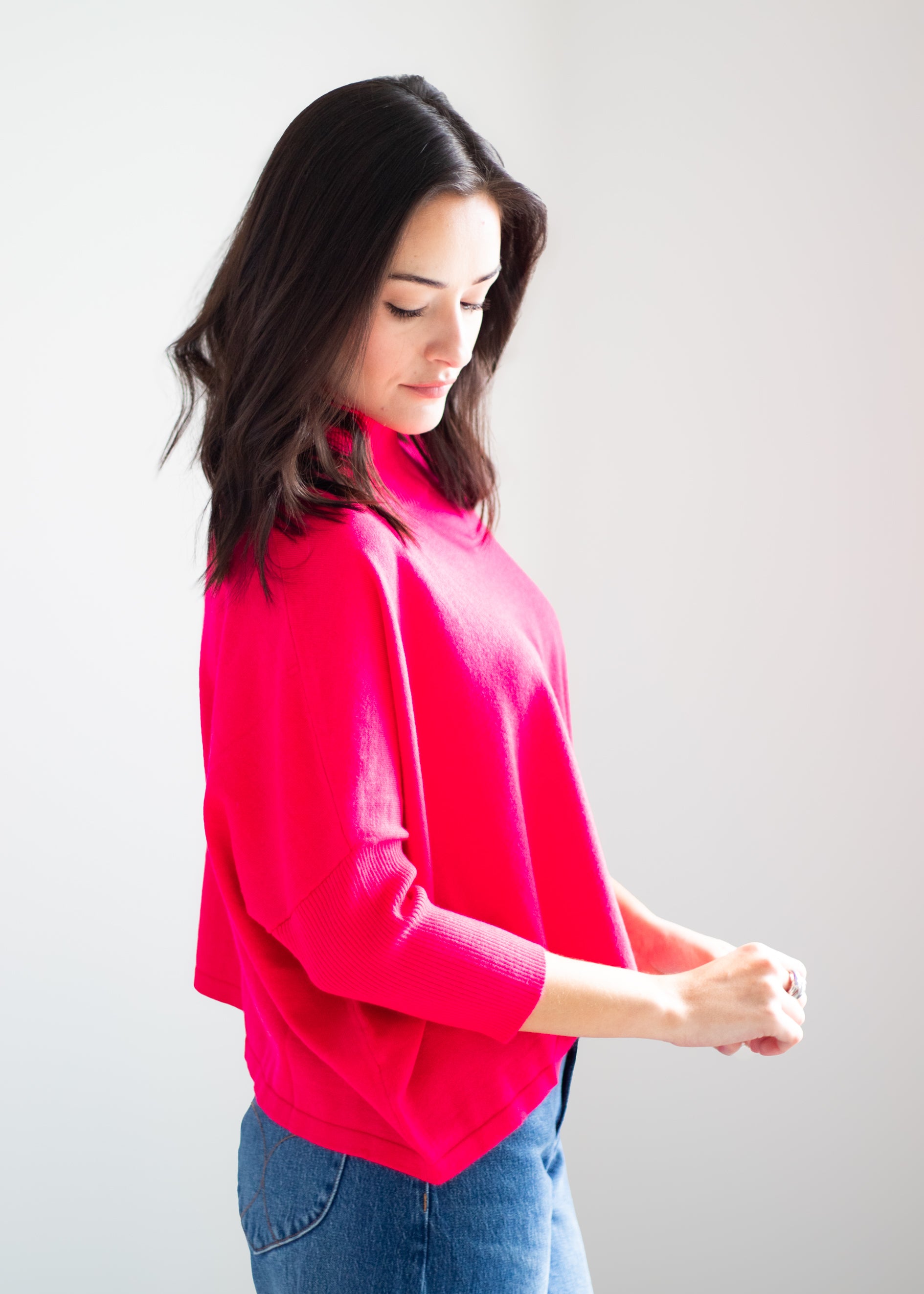 A woman wearing a bright pink knit top with three-quarter sleeves and blue jeans, standing against a plain light gray background.