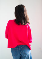 A person viewed from behind wearing a bright neon pink oversized short-sleeve top and blue jeans against a plain light background.
