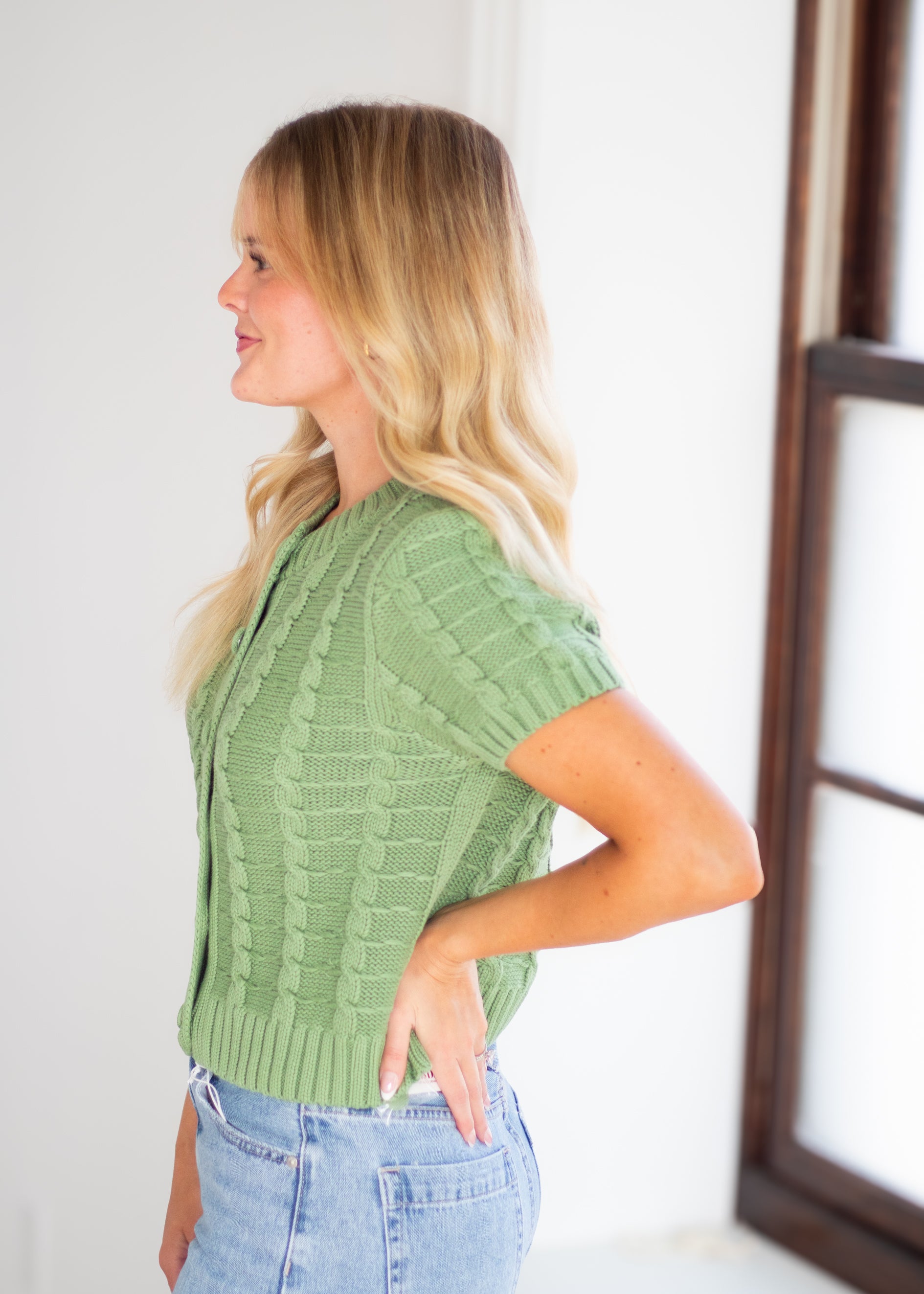 A woman with blonde hair wearing a sage green short-sleeve cable-knit cardigan and light blue jeans, standing in profile in a bright room.