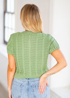 A woman viewed from behind wearing a sage green short-sleeve cable-knit sweater and light blue jeans in a bright indoor setting.