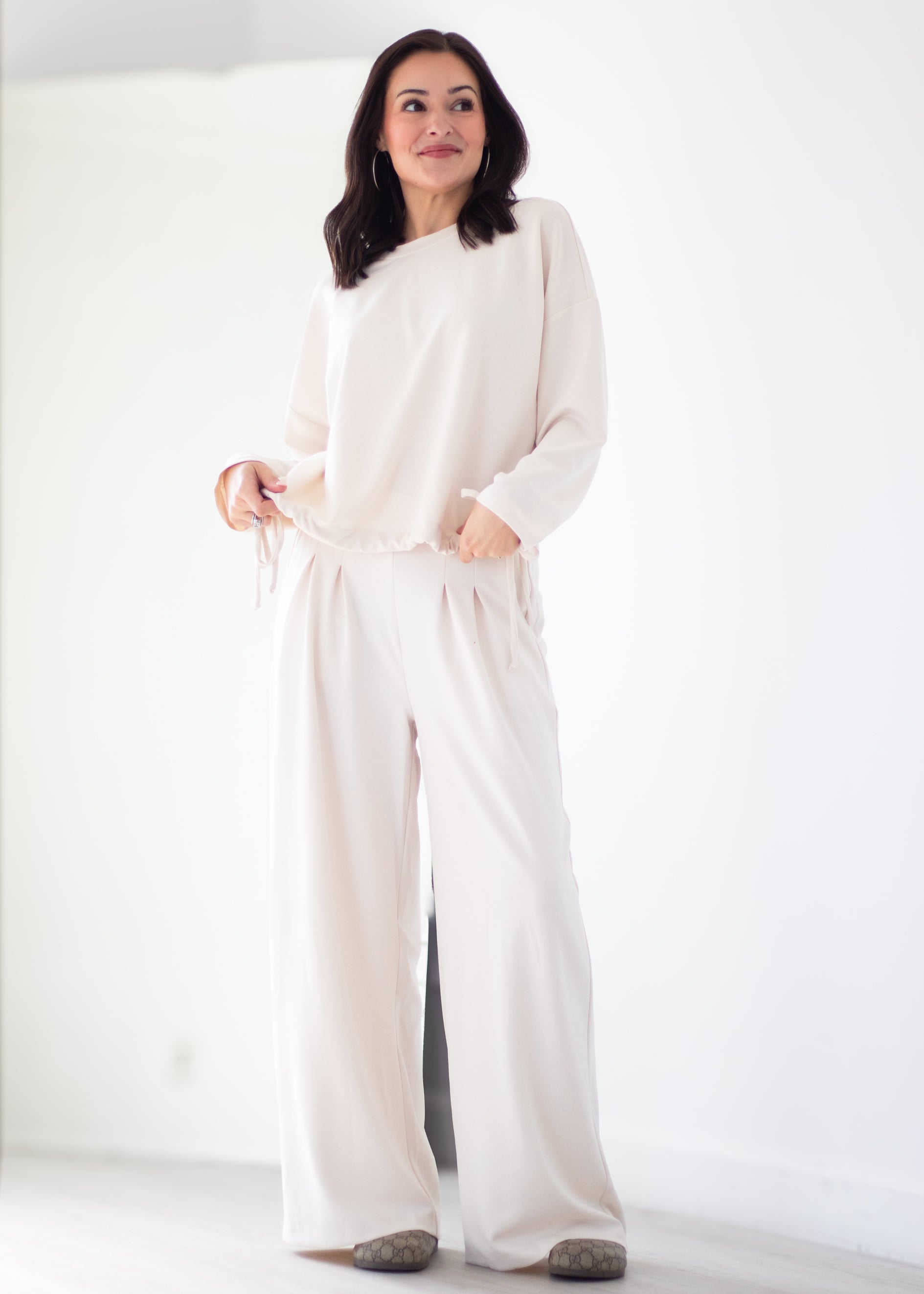 A woman modeling a cream-colored lounge set consisting of a long-sleeve top and matching wide-leg pants, standing in a bright, minimalist white room.