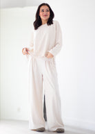 A woman modeling a cream-colored lounge set consisting of a long-sleeve top and matching wide-leg pants, standing in a bright, minimalist white room.