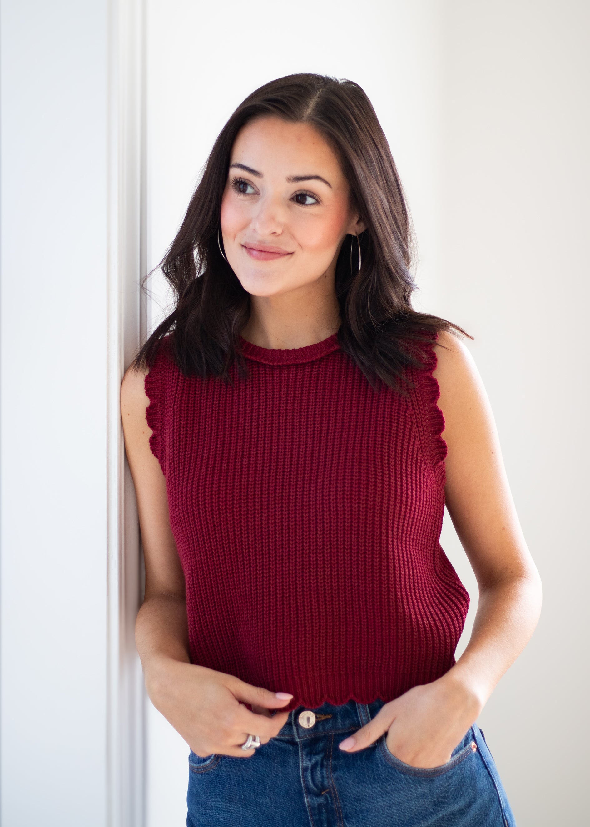 A woman with dark hair wearing a sleeveless, ribbed knit top in deep burgundy with scalloped edges, paired with blue jeans, standing against a white wall.