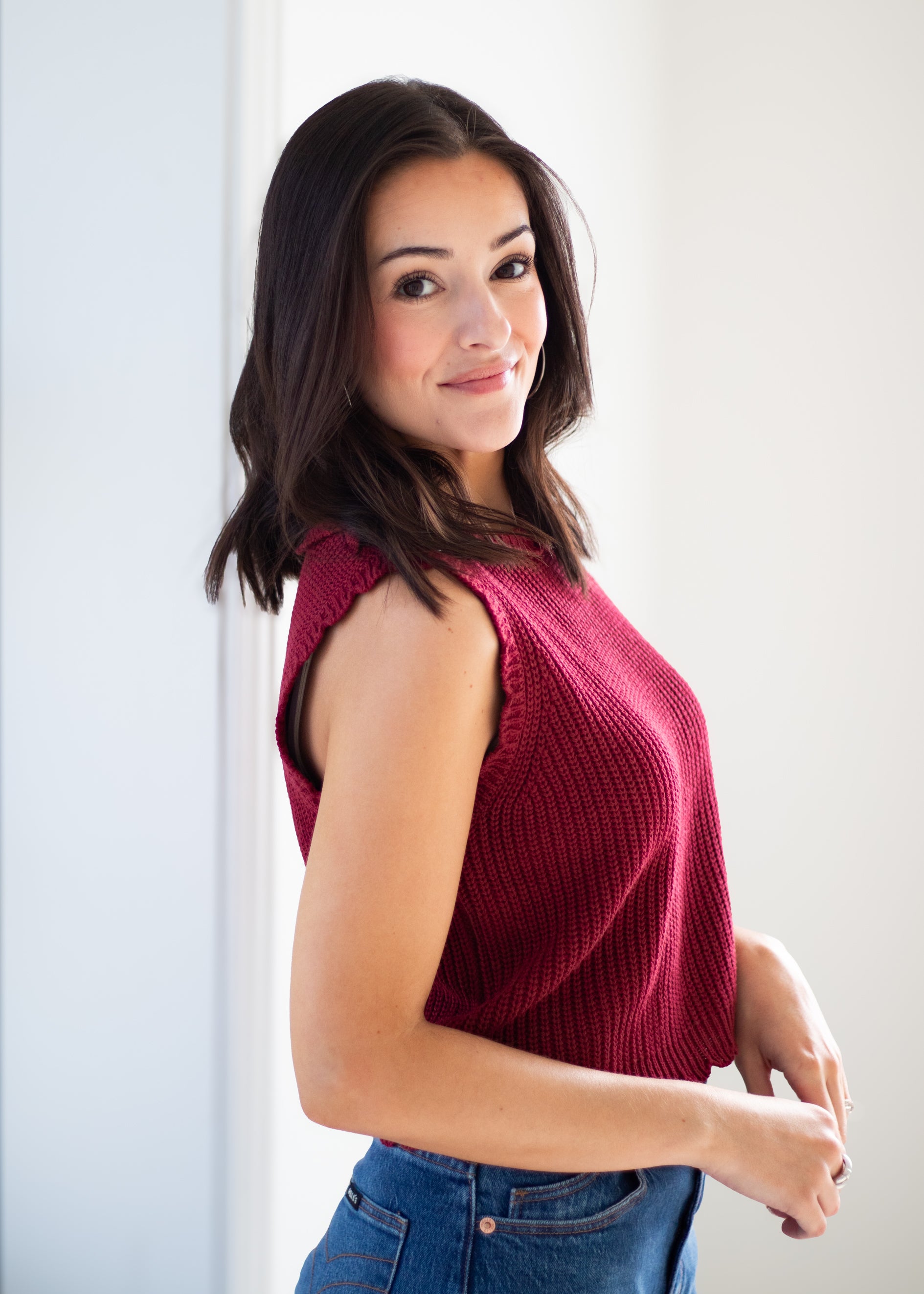 A woman with dark hair wearing a sleeveless, ribbed knit top in deep burgundy and blue jeans, posing in a brightly lit indoor setting.