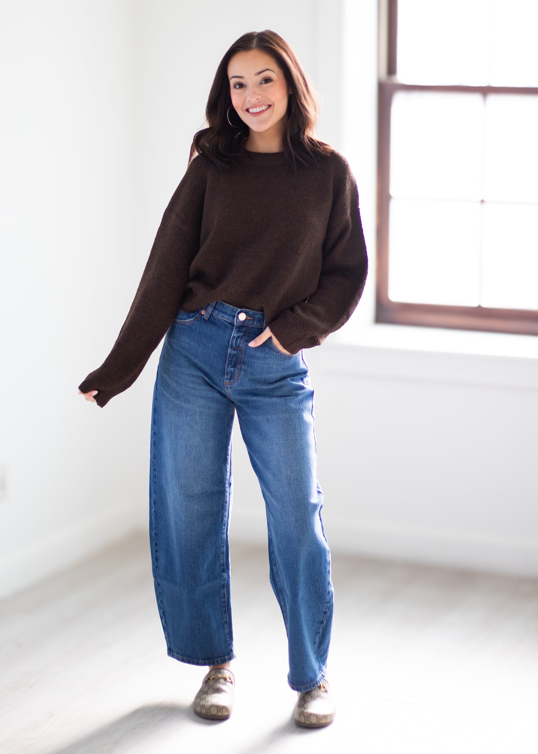 A woman modeling a dark brown long-sleeve knit sweater and wide-leg blue jeans in a bright, minimalist room with a window.