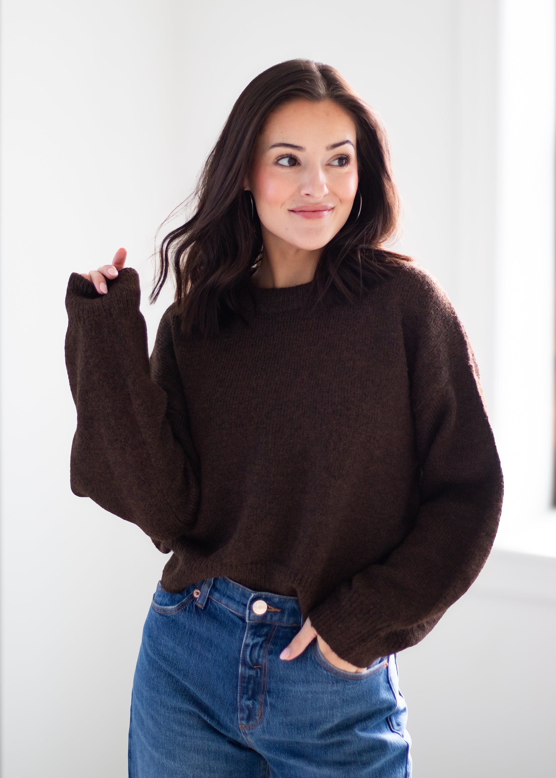 A woman wearing a dark brown fuzzy knit crewneck sweater and blue jeans, standing against a bright white background.
