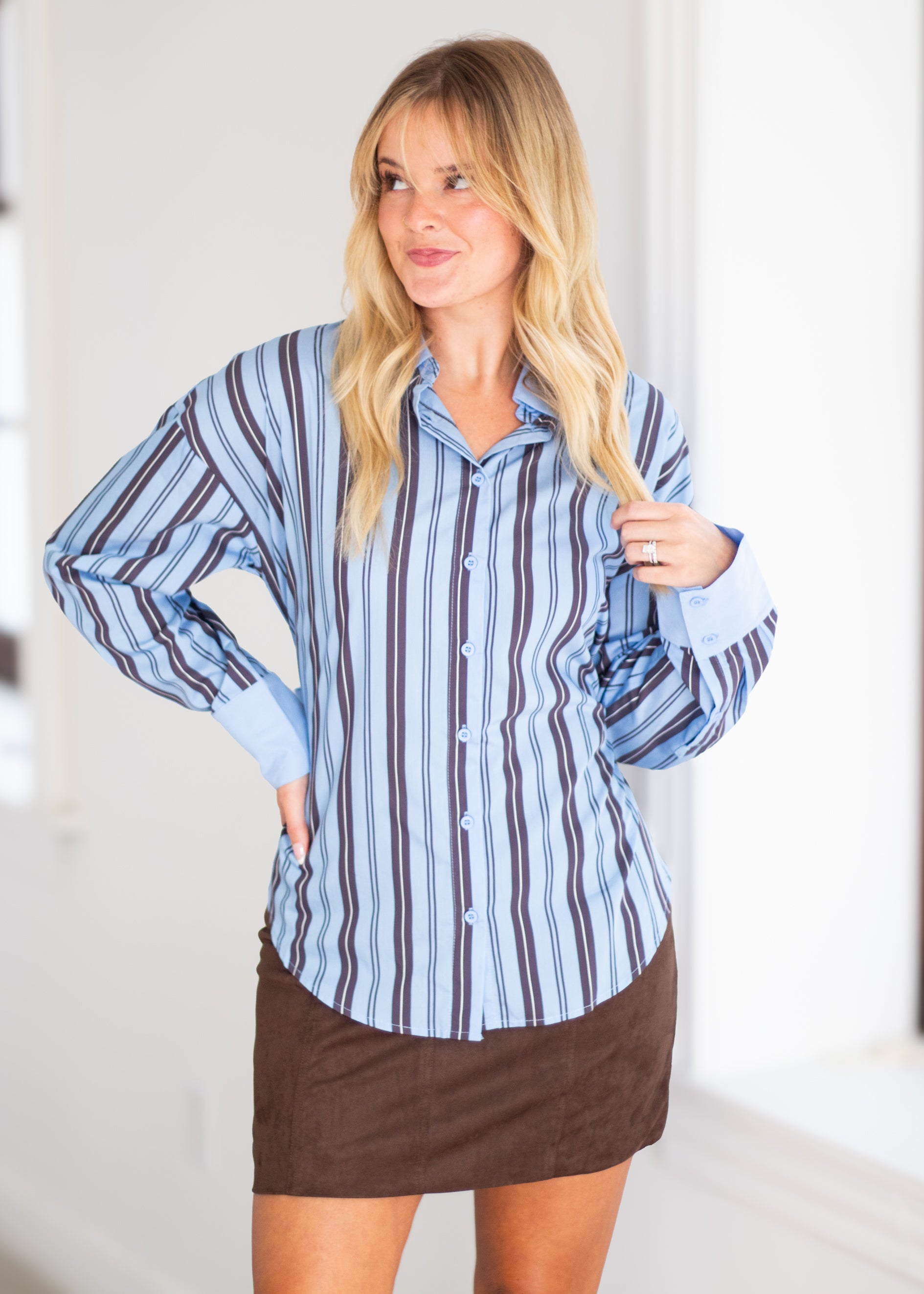 A woman modeling a light blue button-down shirt with vertical dark brown and navy stripes, paired with a brown mini skirt, in a bright indoor setting.