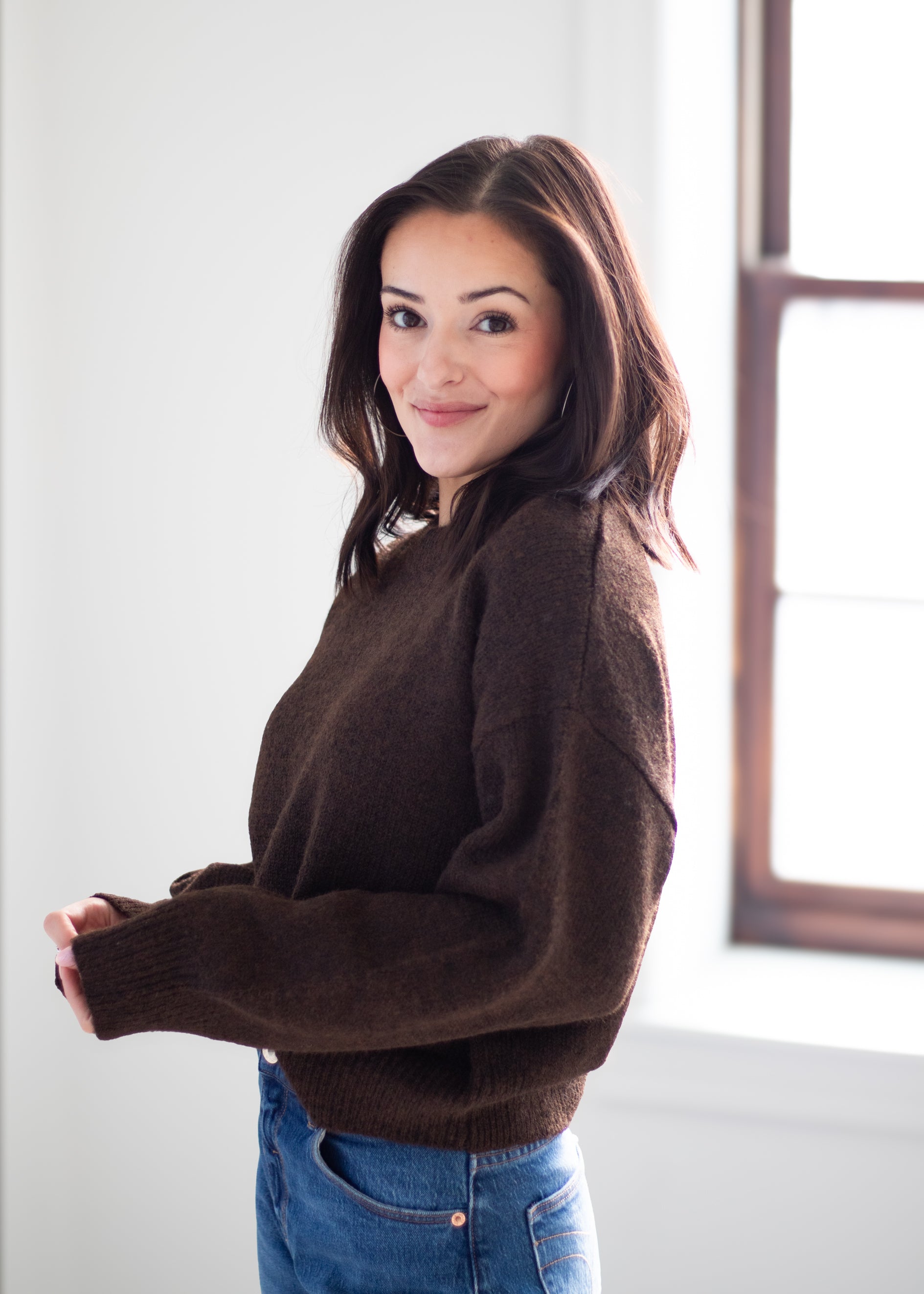 A woman wearing a dark brown knit sweater and blue jeans, standing in a brightly lit room with a window in the background.