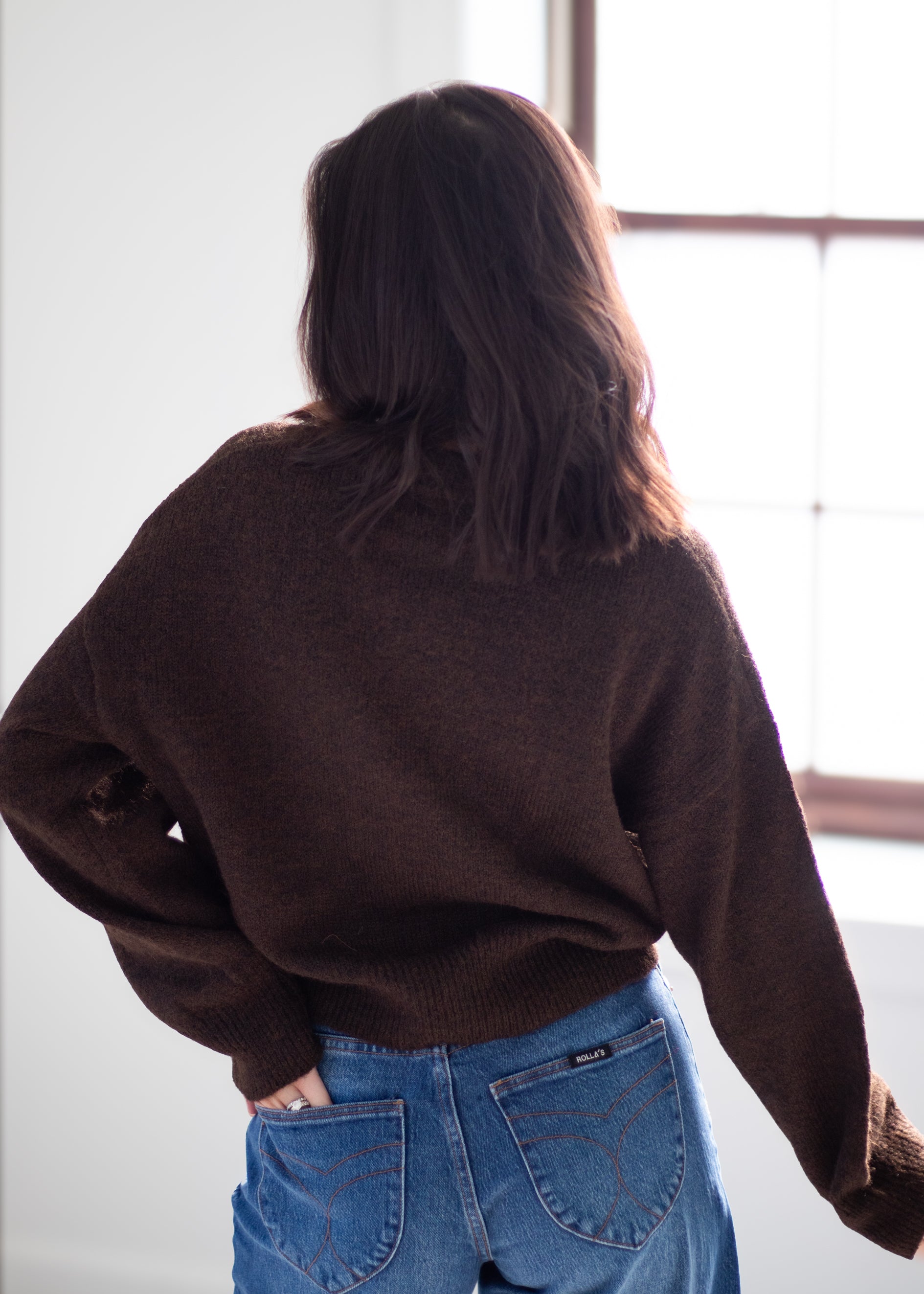 A person viewed from behind wearing a dark brown knit sweater and blue jeans, standing indoors near a bright window.