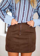 A person wearing a brown mini skirt with a front zipper and a light blue and brown vertically striped button-up shirt, cropped from the chest to the upper thighs.