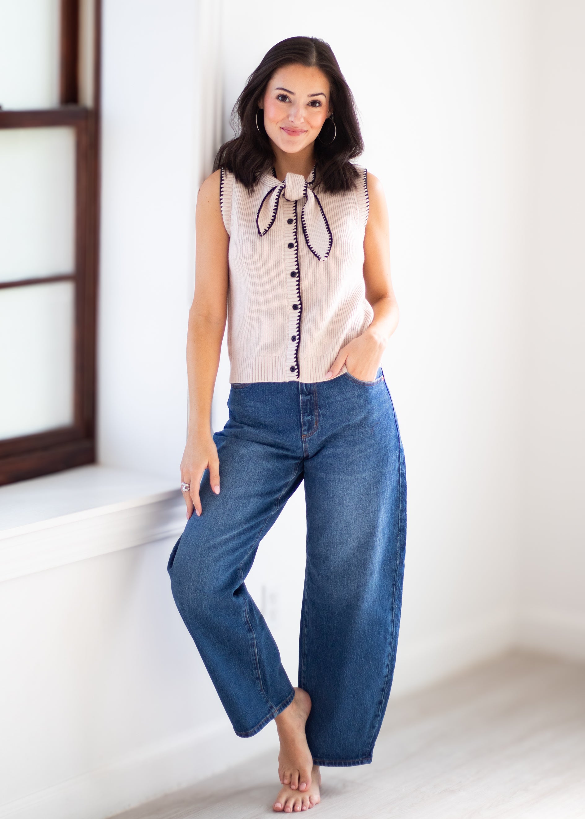 A woman stands barefoot indoors wearing a sleeveless light beige top with a dark bow tie detail and button front, paired with wide-leg blue jeans.