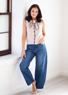 A woman stands barefoot indoors wearing a sleeveless light beige top with a dark bow tie detail and button front, paired with wide-leg blue jeans.