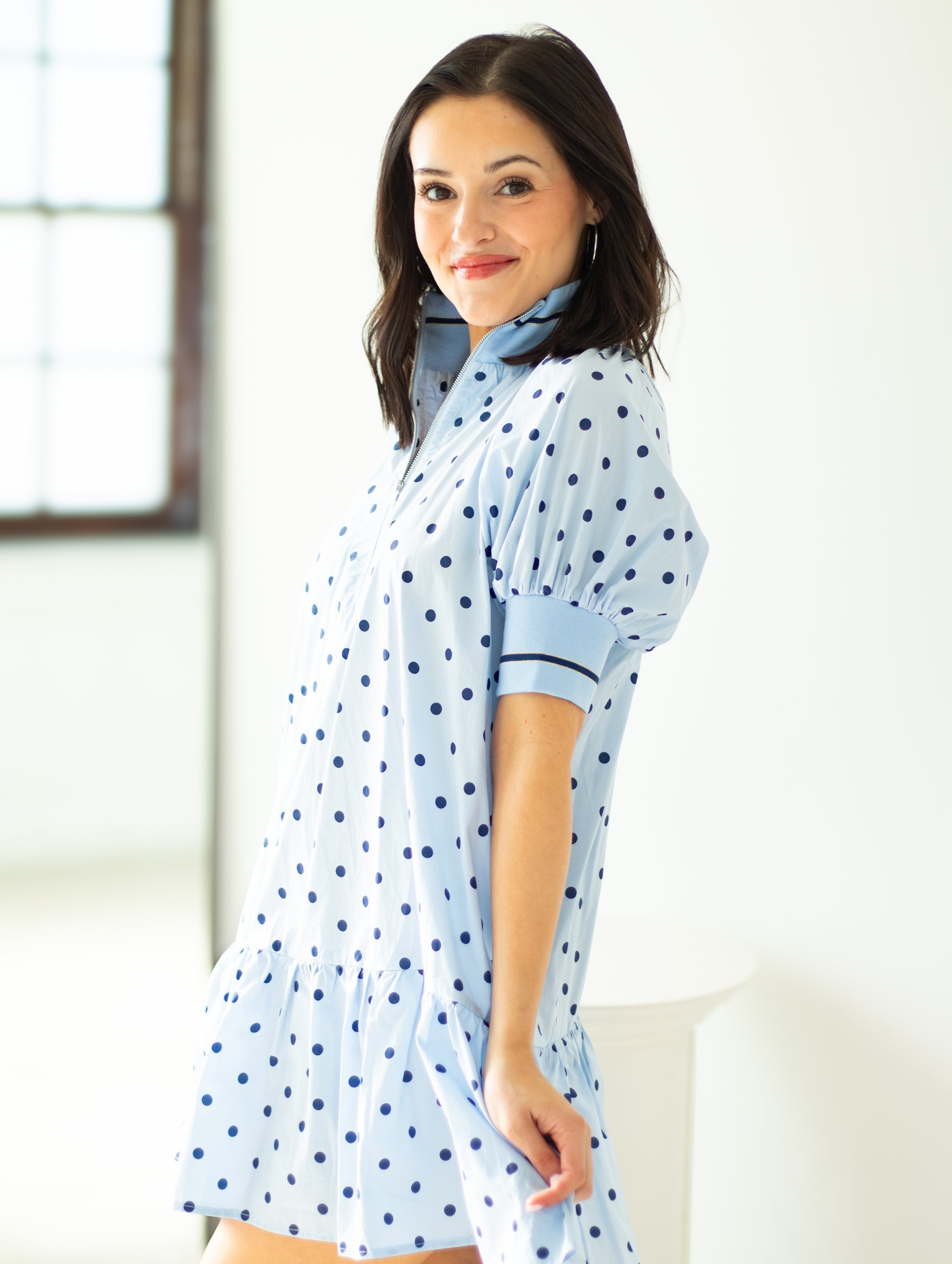 Woman wearing a light blue dress with dark blue polka dots in a bright room.