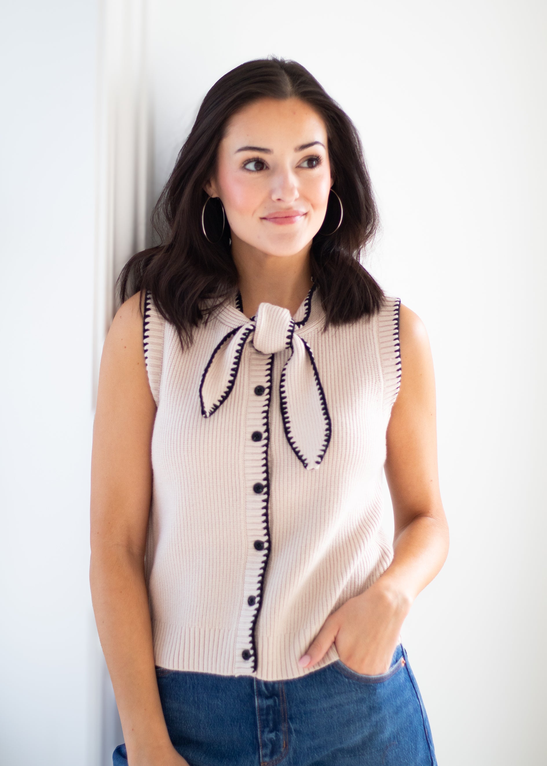 A woman wearing a sleeveless ribbed knit top in a light beige or cream color with black trim and a large black bow detail at the neckline, paired with blue jeans against a white wall.