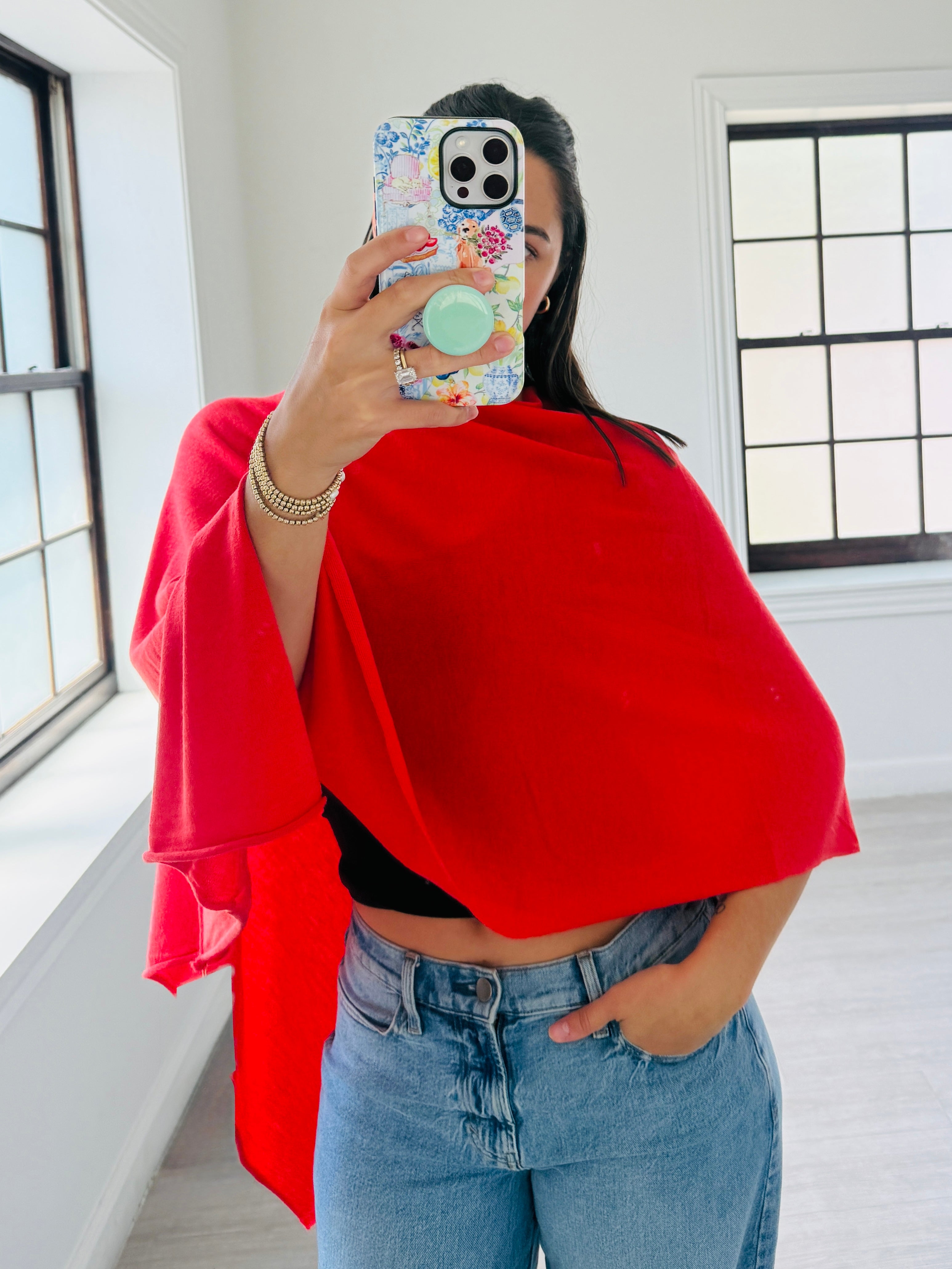 A woman takes a mirror selfie wearing a bright red draped wrap or shawl over a black crop top and blue jeans in a bright room with white walls and windows.
