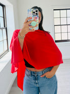 A woman takes a mirror selfie wearing a bright red draped wrap or shawl over a black crop top and blue jeans in a bright room with white walls and windows.