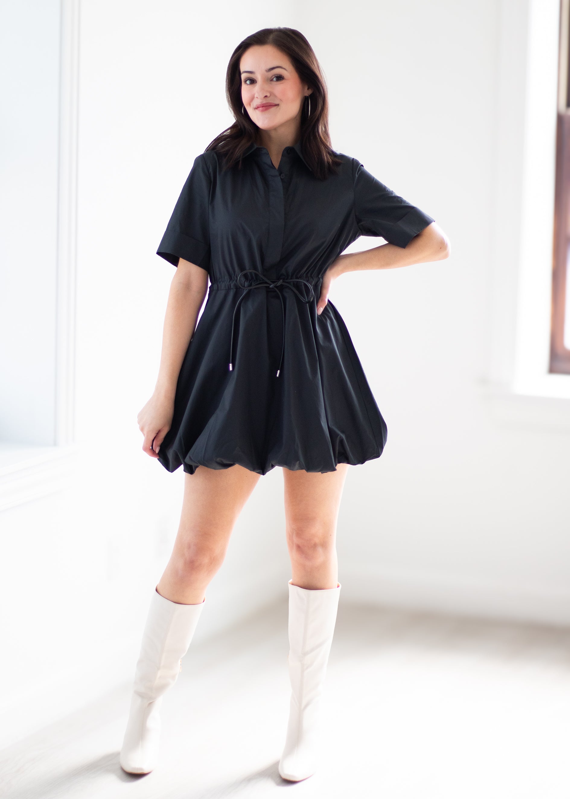 A woman modeling a black short-sleeve shirt dress with a tie waist, paired with white knee-high boots, standing in a bright white room.
