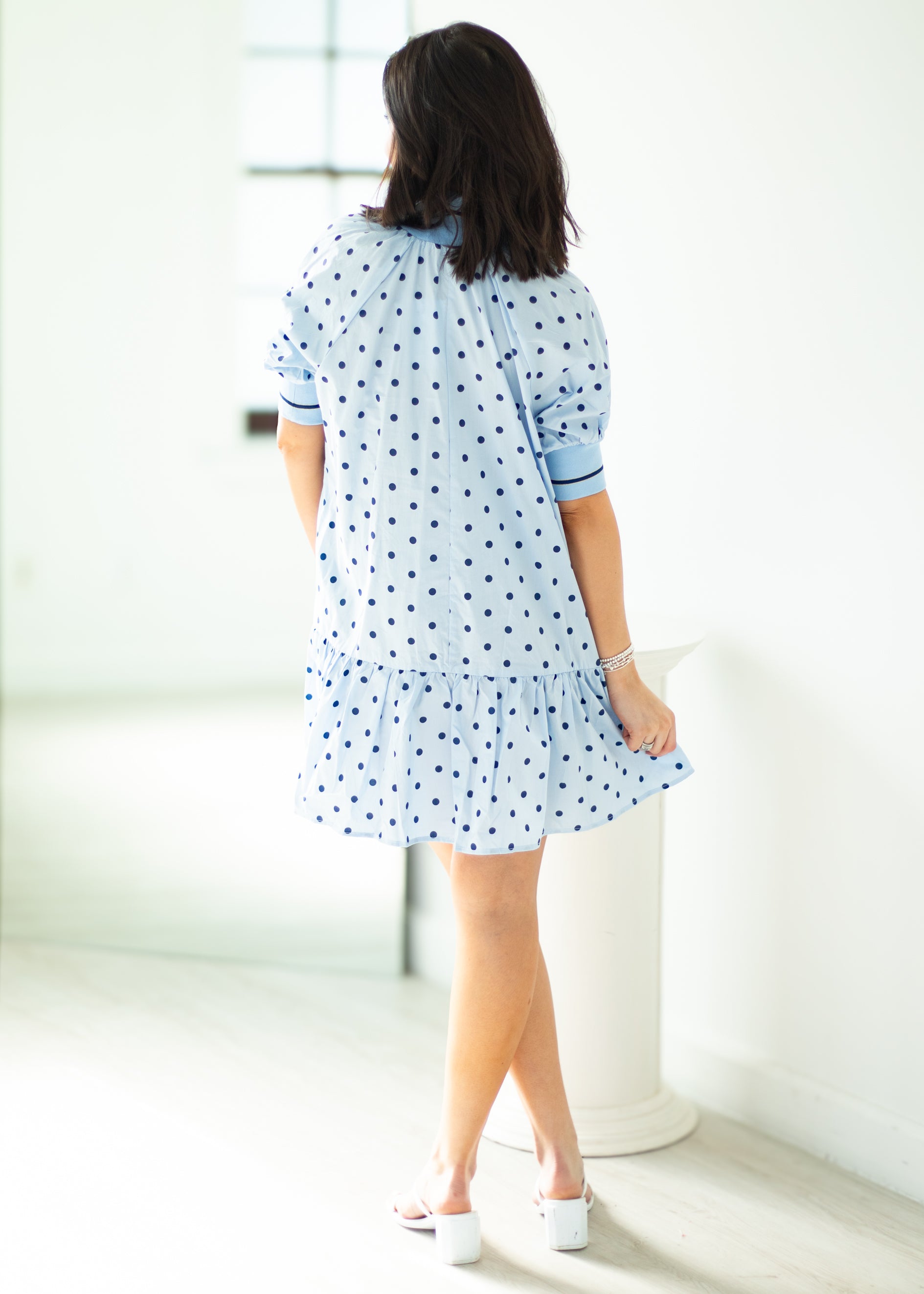 Woman wearing a light blue dress with white polka dots in a minimal indoor setting.