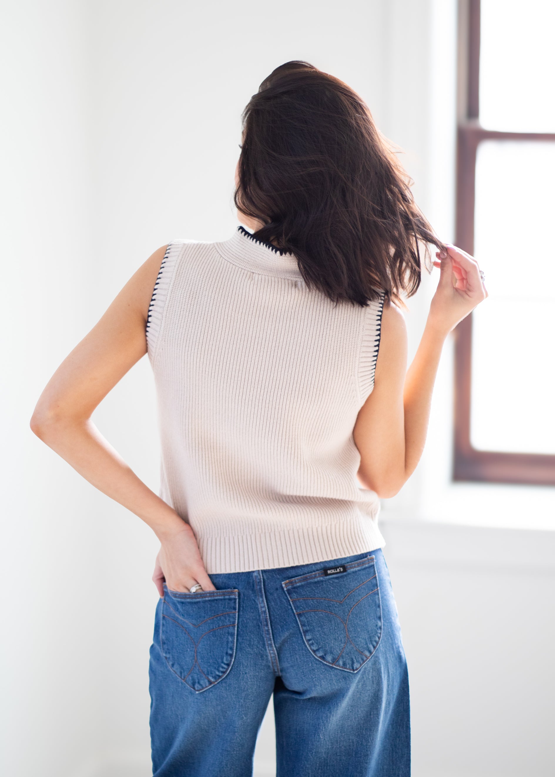 A woman shown from behind wearing a sleeveless ribbed knit sweater vest in a light cream color with black trim, paired with blue jeans, standing in a brightly lit room.