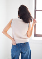 A woman shown from behind wearing a sleeveless ribbed knit sweater vest in a light cream color with black trim, paired with blue jeans, standing in a brightly lit room.