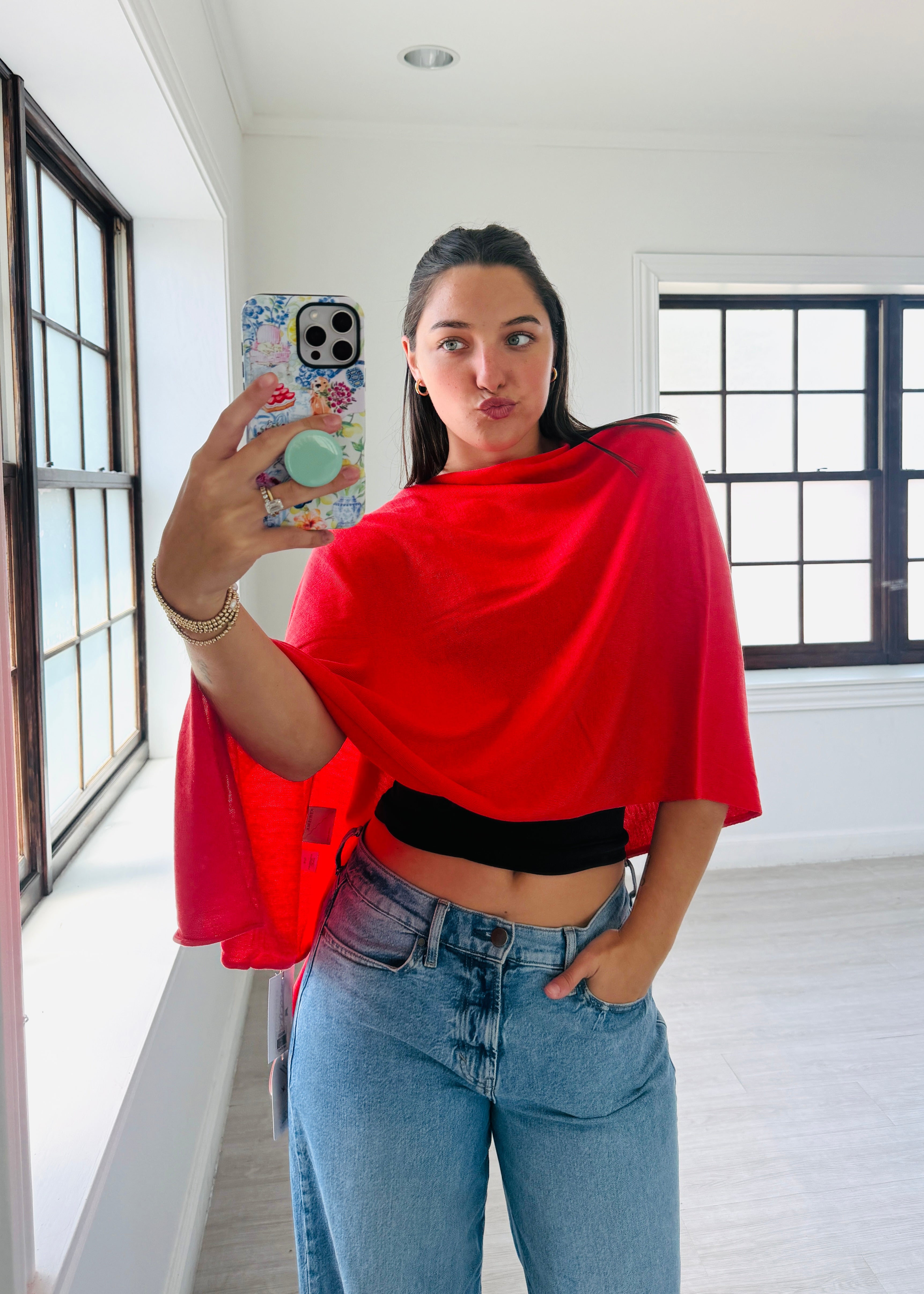 A woman taking a mirror selfie in a bright room, wearing a bright red draped top over a black crop top and light blue jeans.