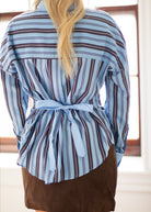 A woman viewed from behind wearing a light blue and dark brown vertically striped long-sleeve shirt with a tie waist, paired with a brown skirt.